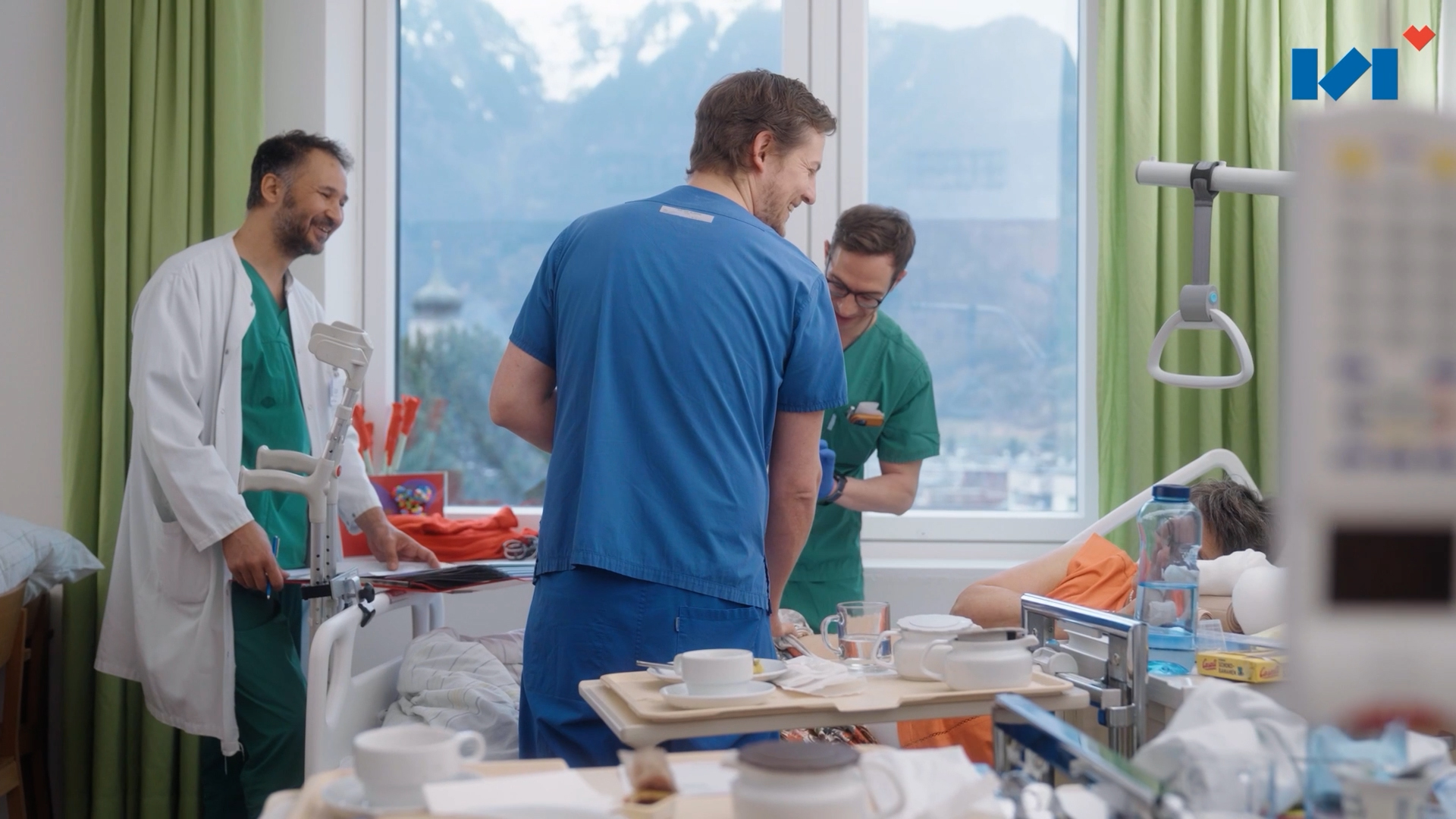 Drei lächelnde Ärzte und Pflegekräfte betreuen eine Patientin im Krankenzimmer des LKH Bludenz; im Hintergrund ist ein Fenster mit Bergpanorama zu sehen.